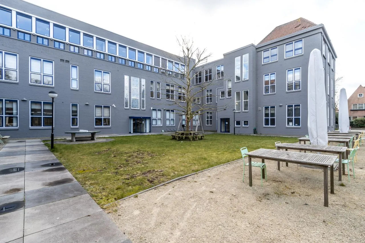 Binnenplaats van een grijs gebouw aan de Lage Naarderweg met zitbanken, tafels en een boom in het midden.